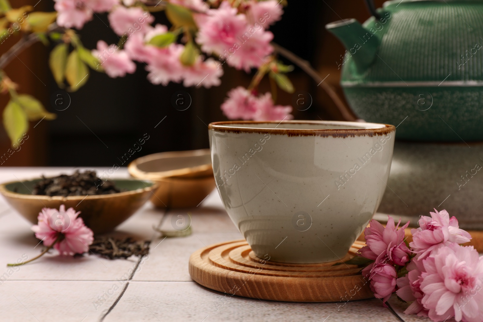 Photo of Traditional ceremony. Cup of brewed tea, teapot and sakura flowers on tiled table, closeup