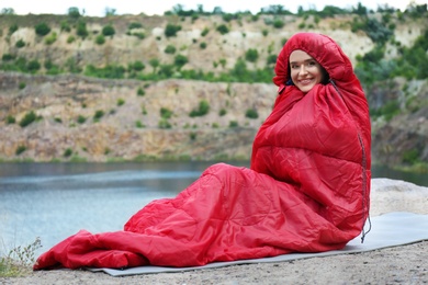 Image of Young camper in sleeping bag sitting on cliff near lake