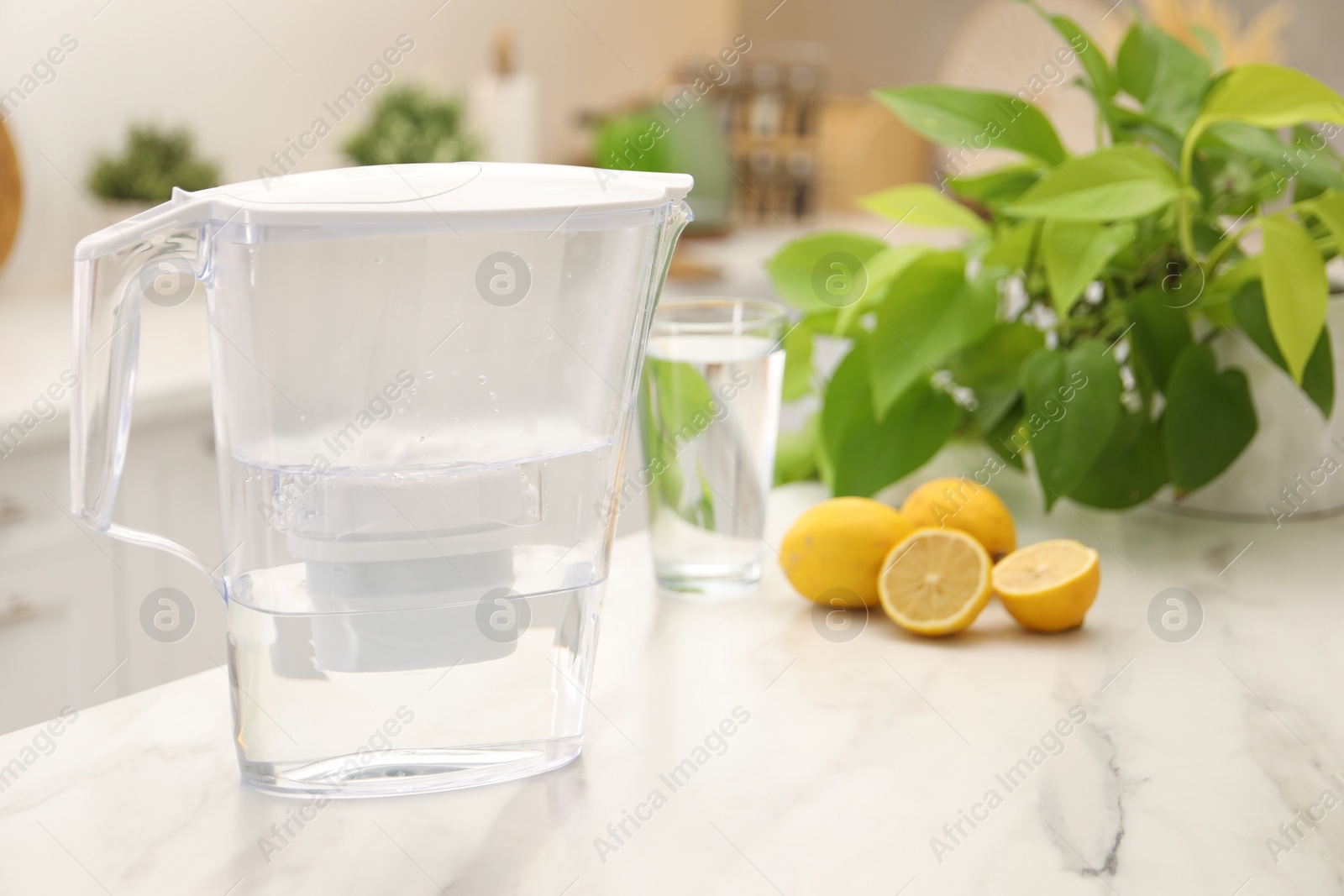 Photo of Water filter jug, glass and lemons on white marble table in kitchen