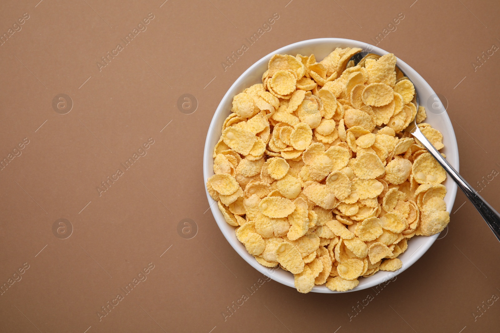 Photo of Breakfast cereal. Tasty corn flakes in bowl and spoon on brown table, top view. Space for text