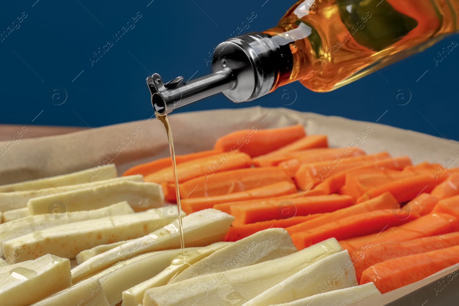 Photo of Pouring oil onto baking tray with parsnips and carrots against blue background, closeup