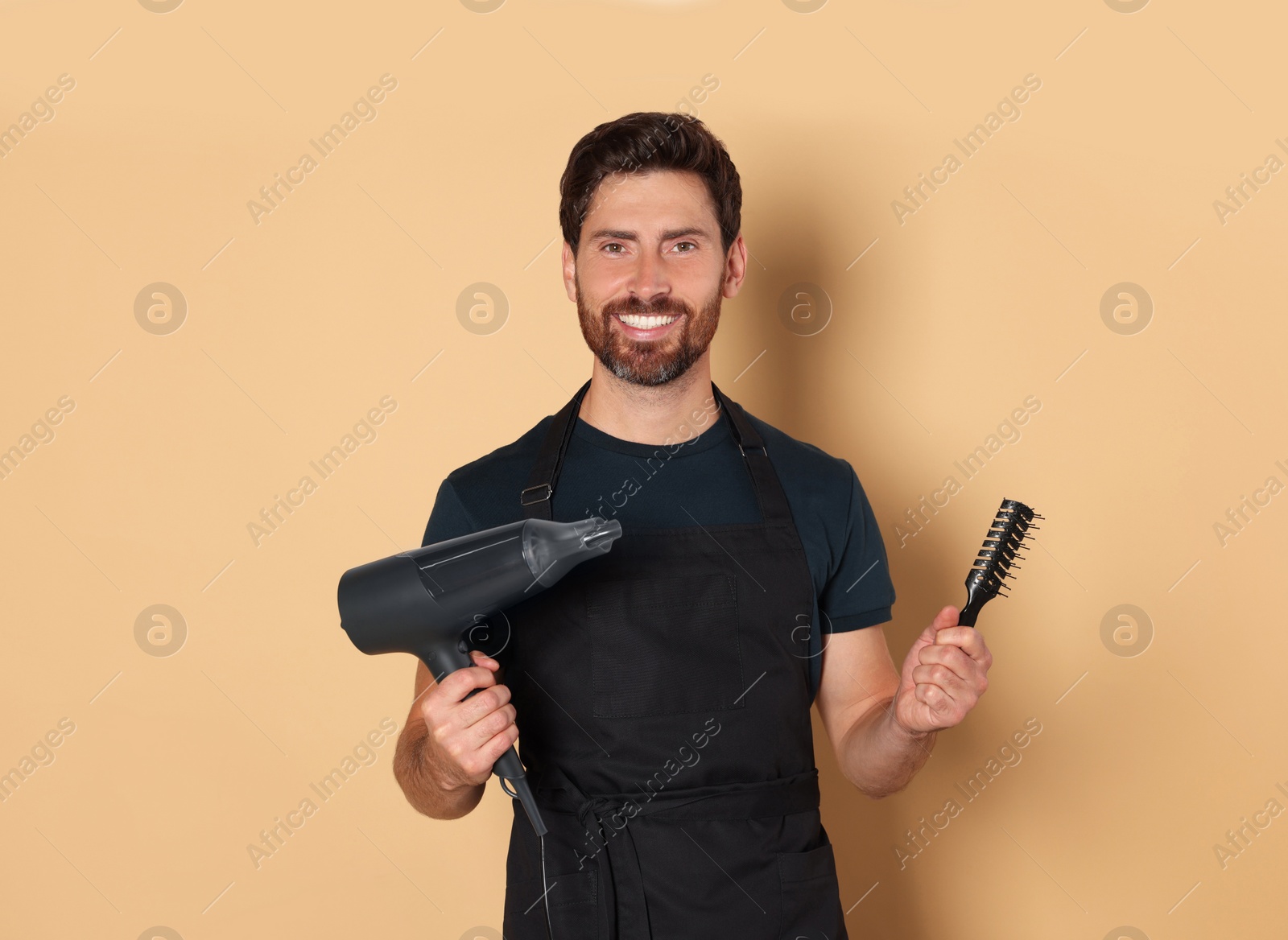 Photo of Smiling hairdresser in apron holding dryer and brush on light brown background