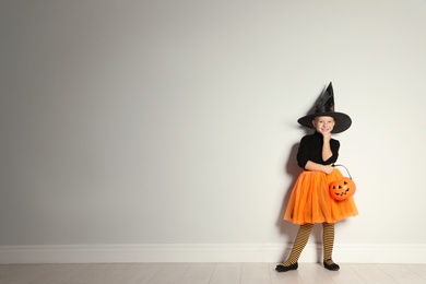 Photo of Cute little girl with pumpkin candy bucket wearing Halloween costume near light wall. Space for text