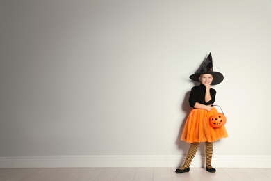 Cute little girl with pumpkin candy bucket wearing Halloween costume near light wall. Space for text