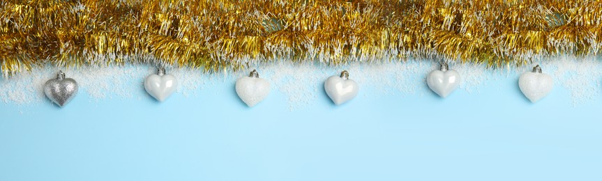 Image of Shiny golden tinsel, Christmas baubles and snow on light blue background, flat lay. Banner design