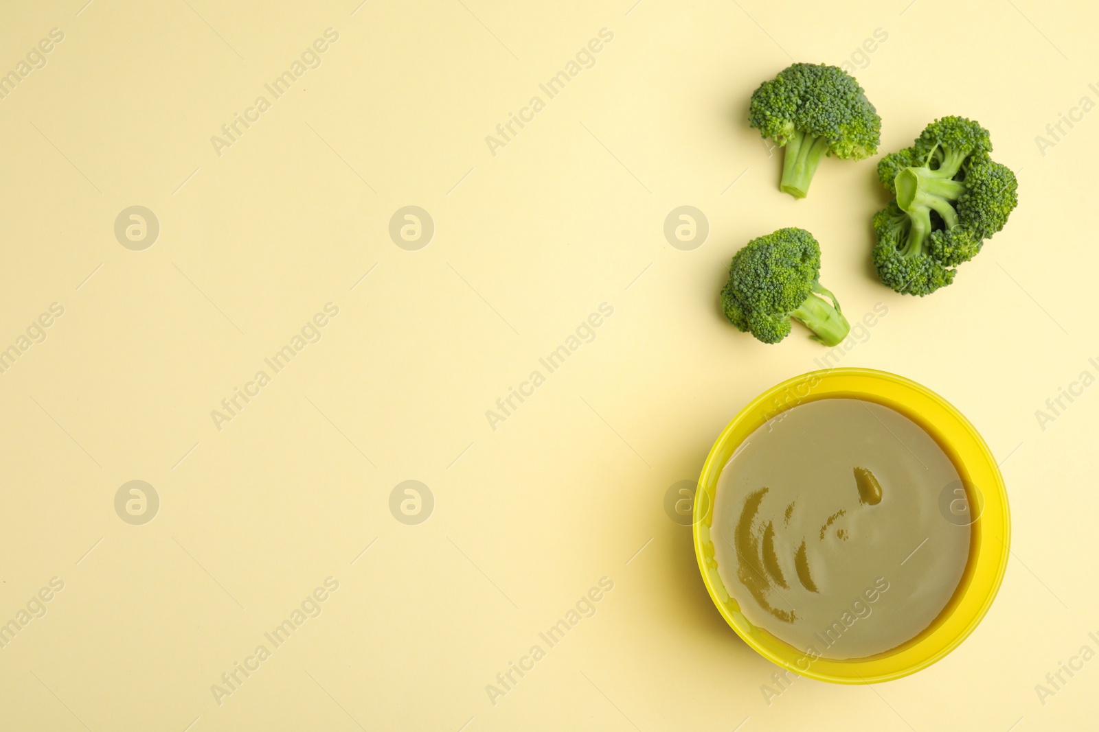 Photo of Baby food in bowl and fresh broccoli on yellow background, flat lay. Space for text