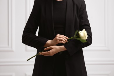 Woman with rose flower near white wall, closeup. Funeral ceremony