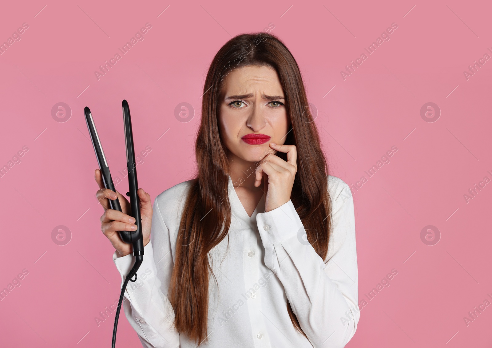 Photo of Upset young woman with flattening iron on light pink background. Hair damage