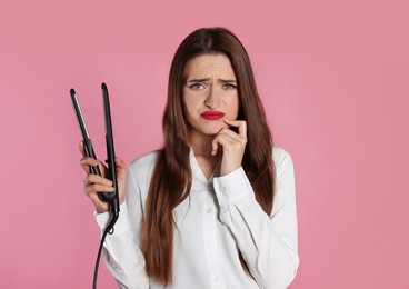 Photo of Upset young woman with flattening iron on light pink background. Hair damage