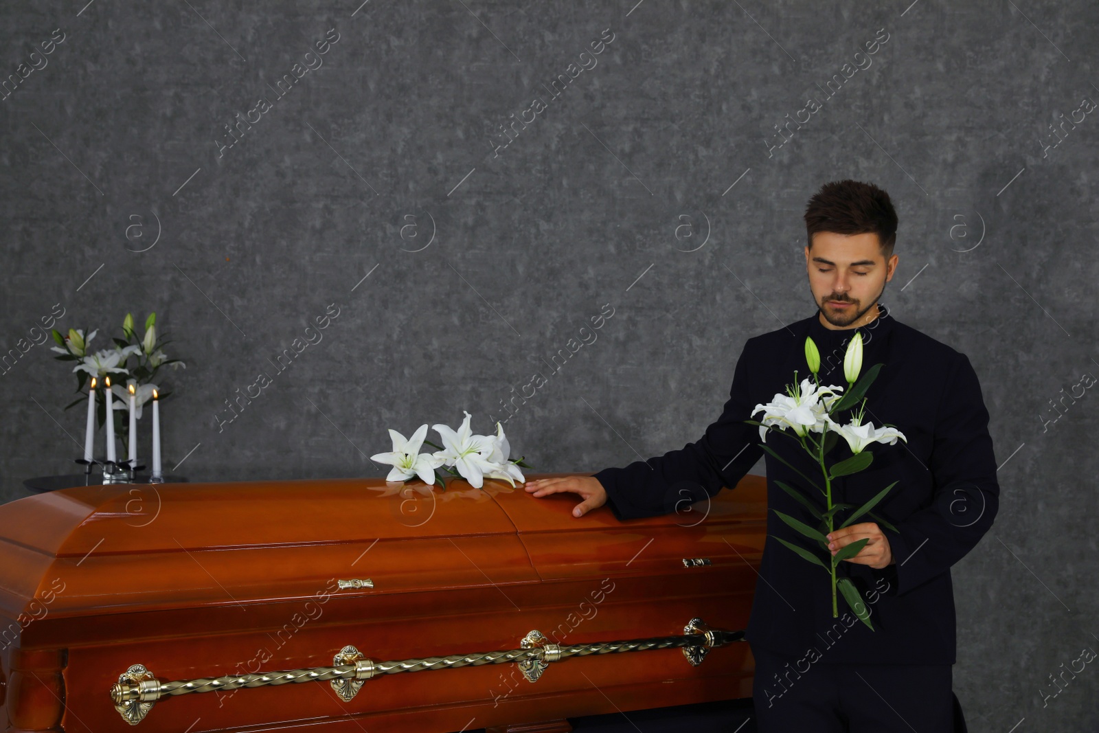 Photo of Sad young man with white lilies near casket in funeral home