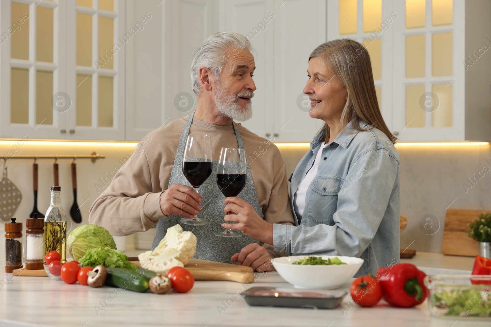 Photo of Happy senior couple with glasses of wine cooking together in kitchen
