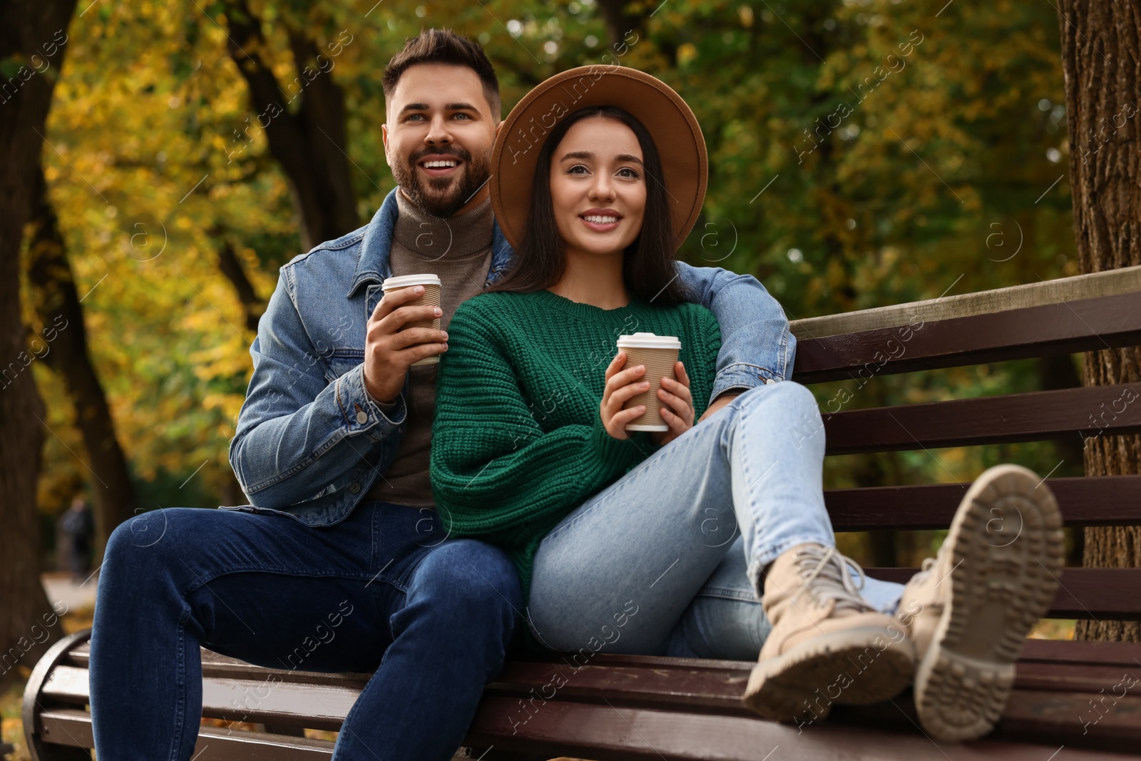 Photo of Happy young couple with cups of coffee spending time together on wooden bench in autumn park