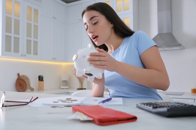 Photo of Young woman with piggy bank at table in kitchen. Money savings