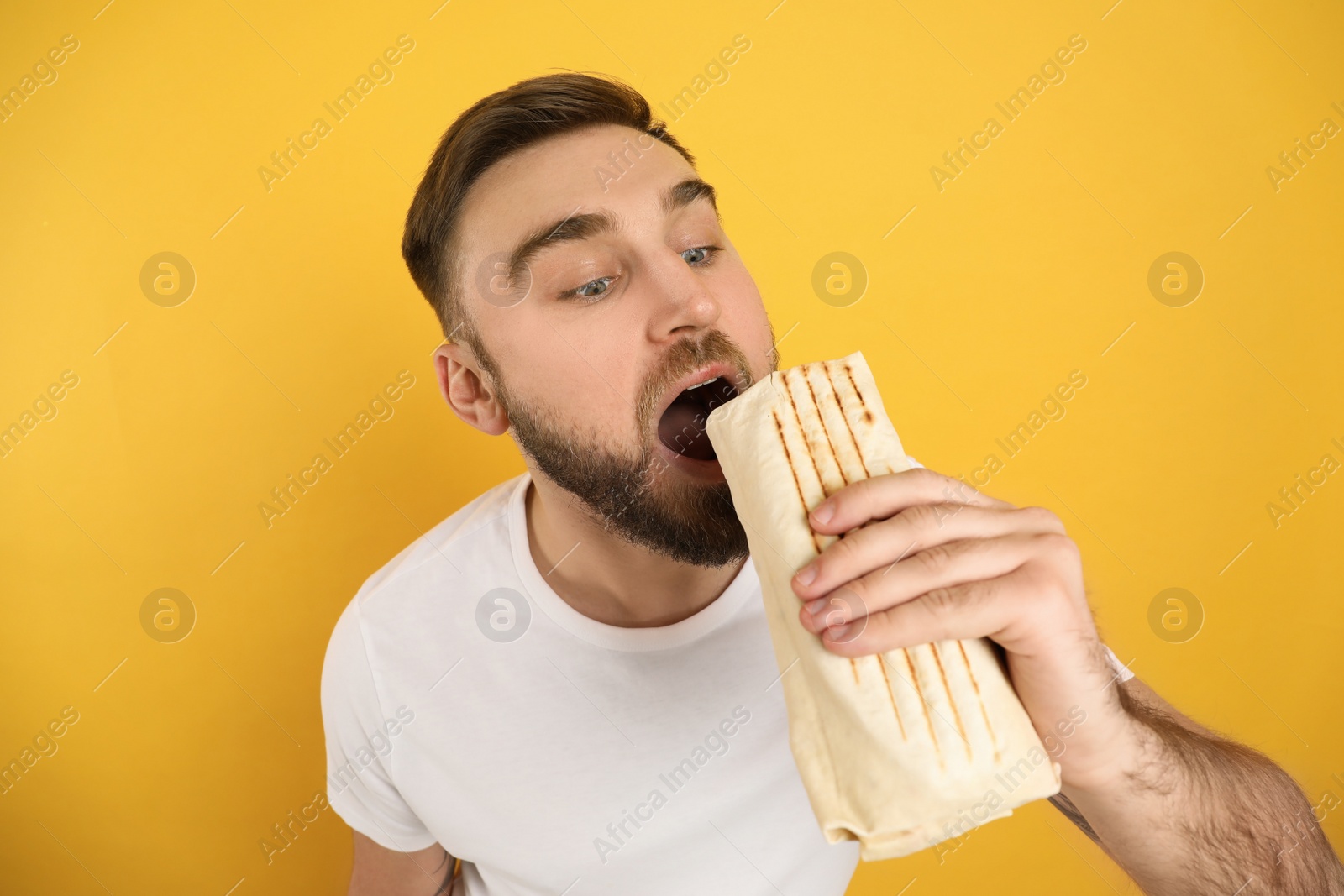 Photo of Young man eating delicious shawarma on yellow background