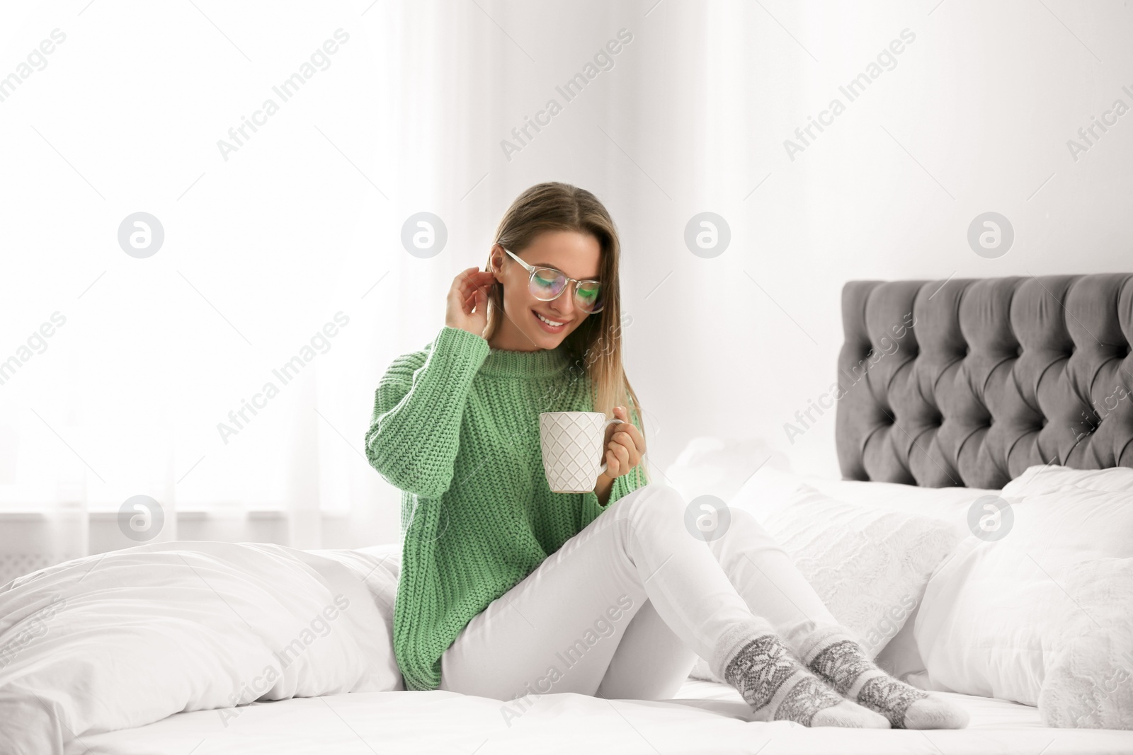 Photo of Young woman in warm sweater with hot drink on bed at home