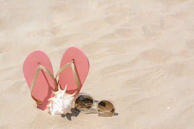 Photo of Stylish pink flip flops, sunglasses and seashell on sandy beach, space for text
