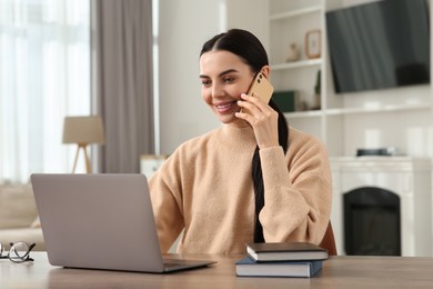 Photo of Happy woman talking on smartphone while working with laptop at desk in room