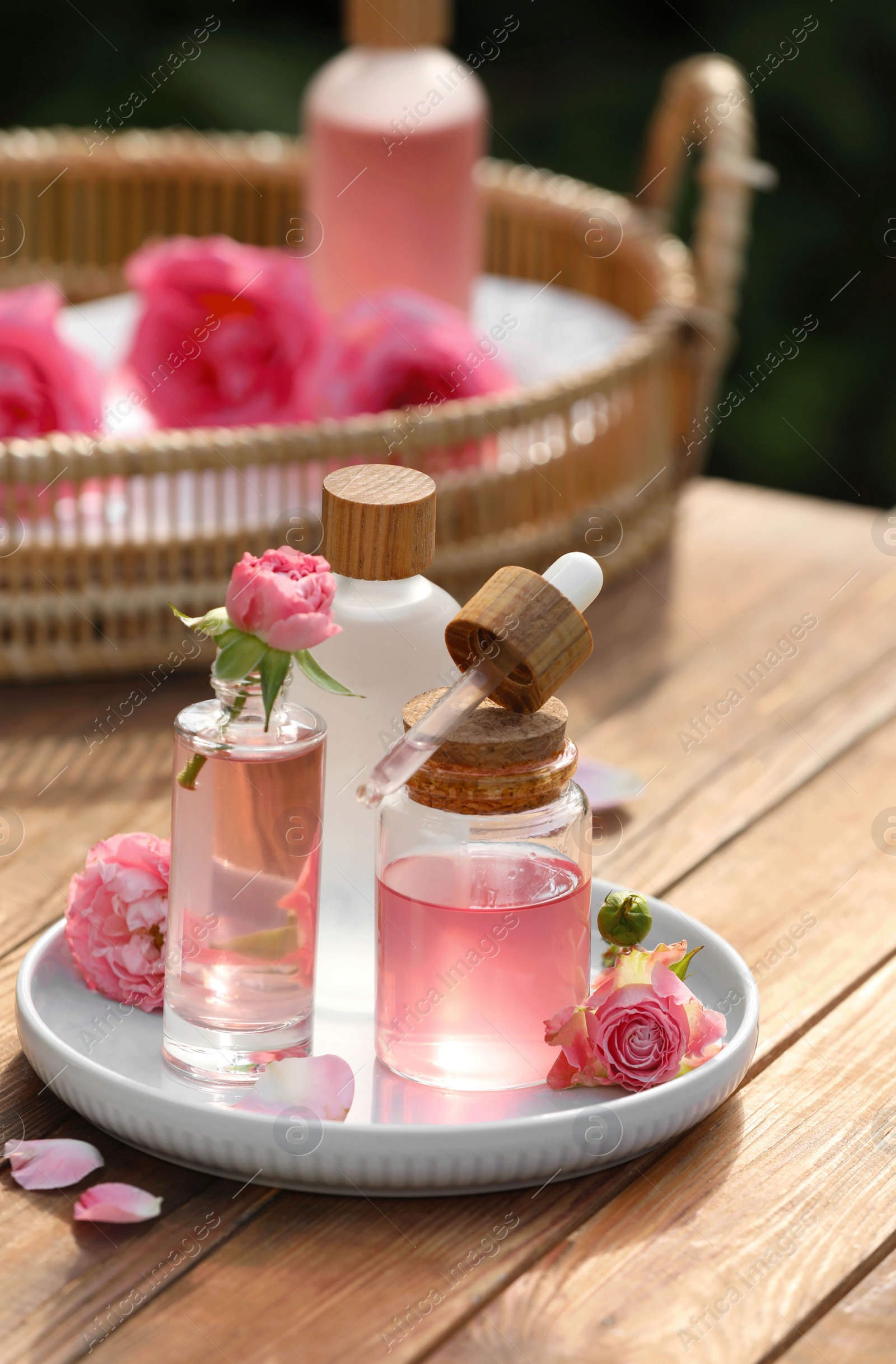 Photo of Bottles of rose essential oil and flowers on wooden table outdoors