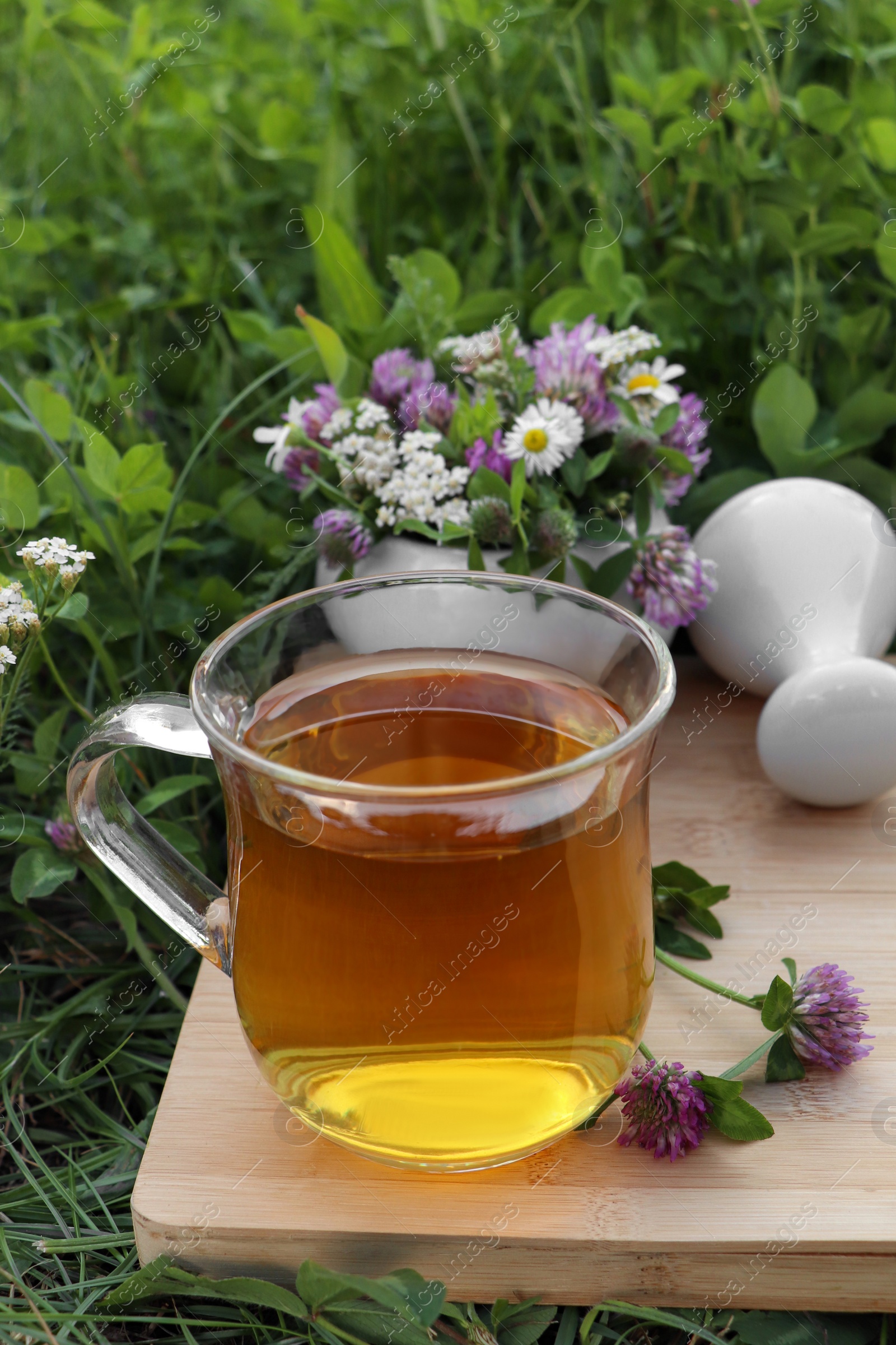 Photo of Cup of aromatic herbal tea, pestle and ceramic mortar with different wildflowers on wooden board in meadow