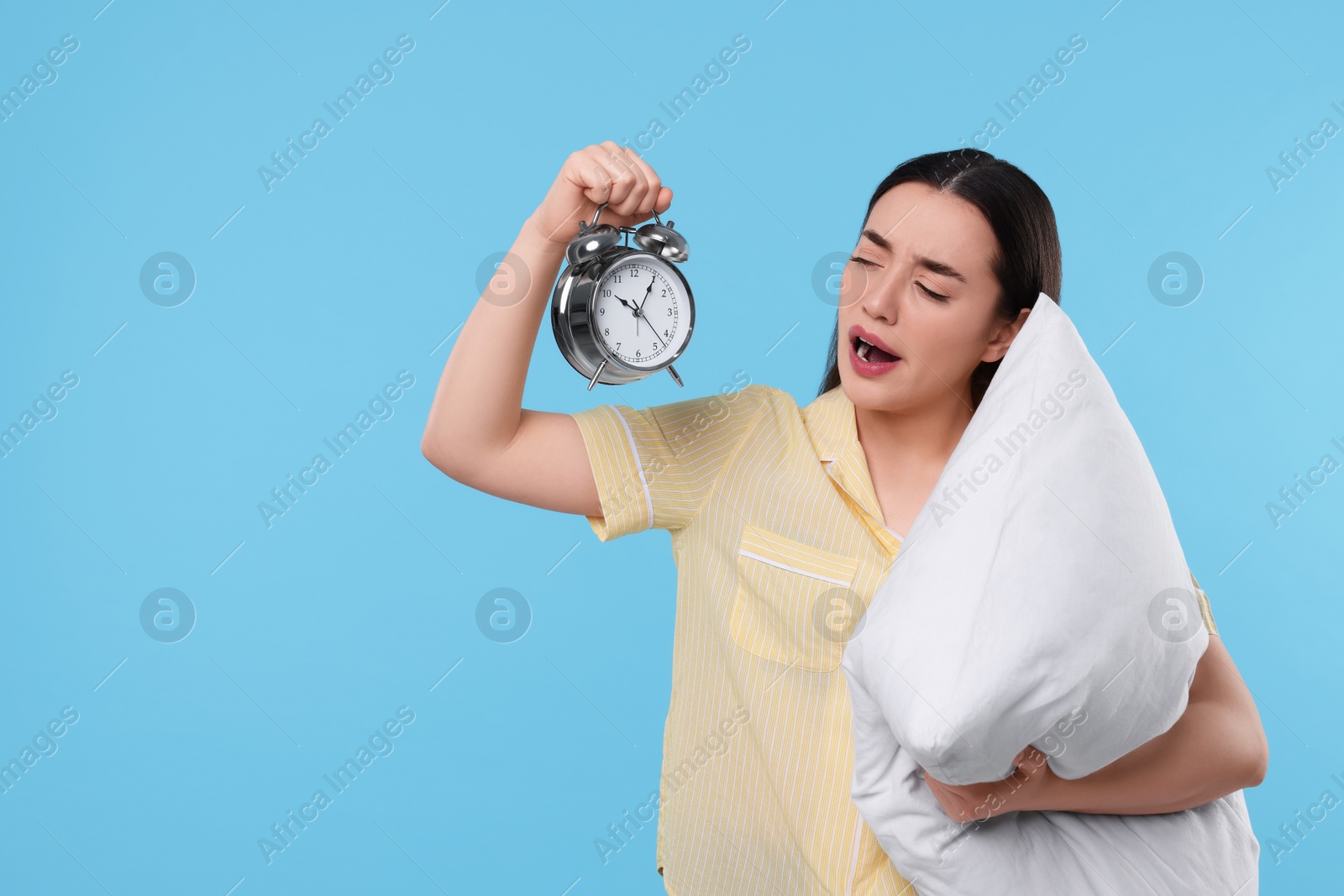 Photo of Tired young woman with pillow and alarm clock yawning on light blue background, space for text. Insomnia problem