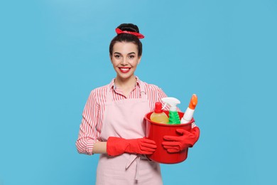 Young housewife holding bucket with cleaning supplies on light blue background