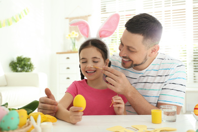 Happy daughter with bunny ears headband and her father having fun while painting Easter egg at home