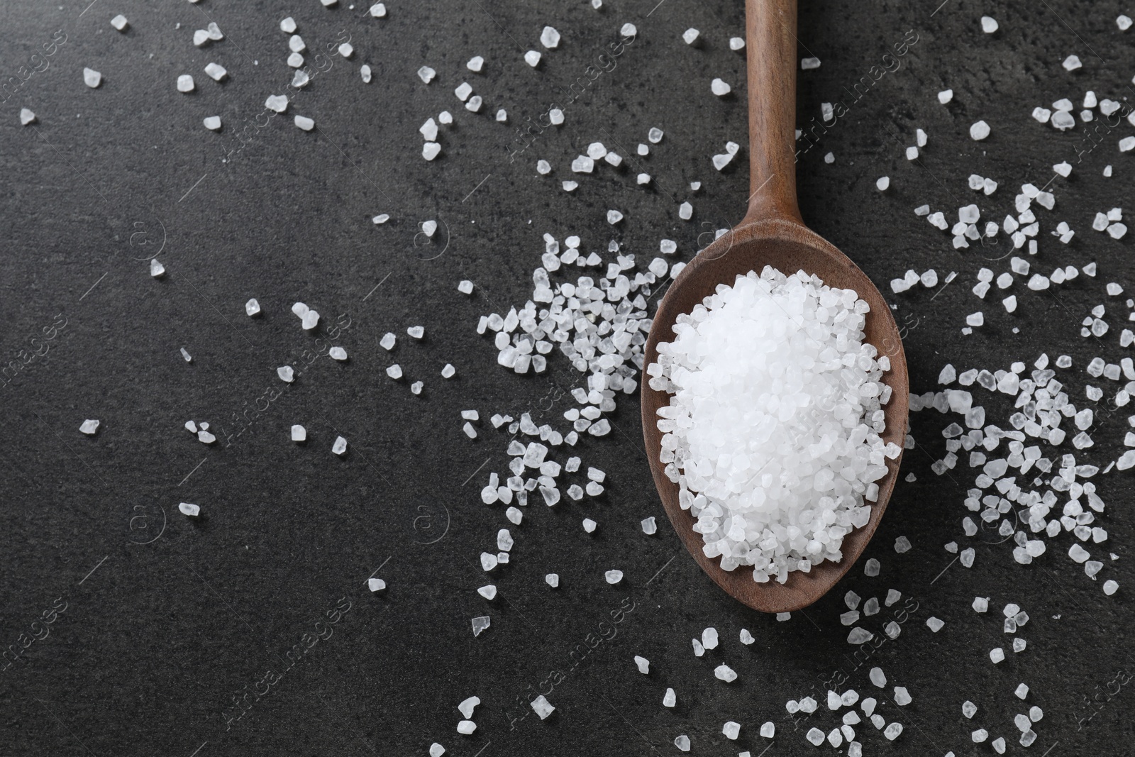 Photo of Natural salt and wooden spoon on black table, top view