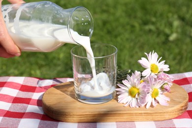 Photo of Woman pouring fresh milk from bottle into glass on checkered blanket outdoors, closeup
