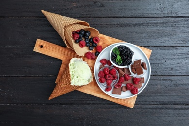 Photo of Delicious ice cream in wafer cone, chocolate and berries on black wooden table, top view