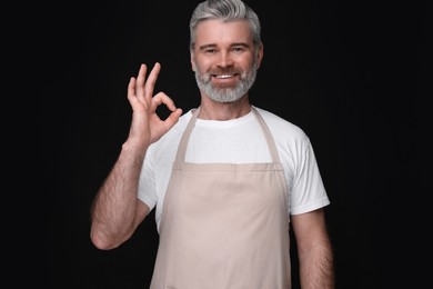 Photo of Happy man in kitchen apron showing OK gesture on black background. Mockup for design