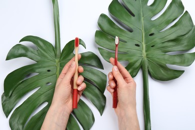 Photo of Woman holding natural bamboo and plastic toothbrushes above tropical leaves on white background, top view