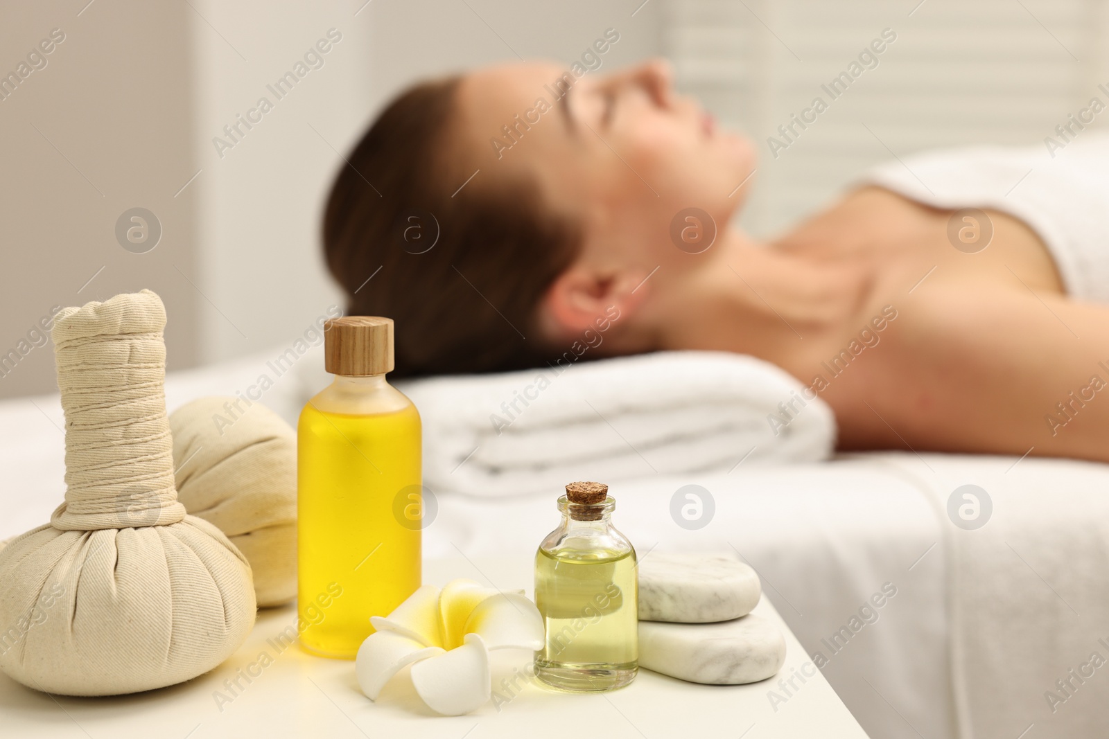 Photo of Aromatherapy. Woman relaxing on massage couch in spa salon, focus on bottles of essential oils, herbal bags, stones and plumeria flower