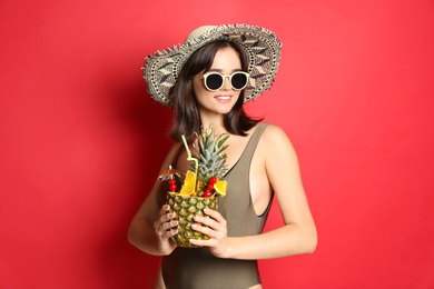 Beautiful woman in stylish swimsuit holding tropical cocktail on red background