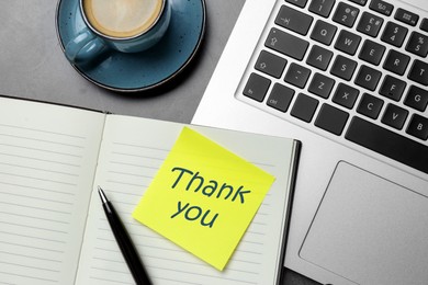 Image of Sticky note with phrase Thank You, notebook, laptop, pen and cup of coffee on grey table, flat lay