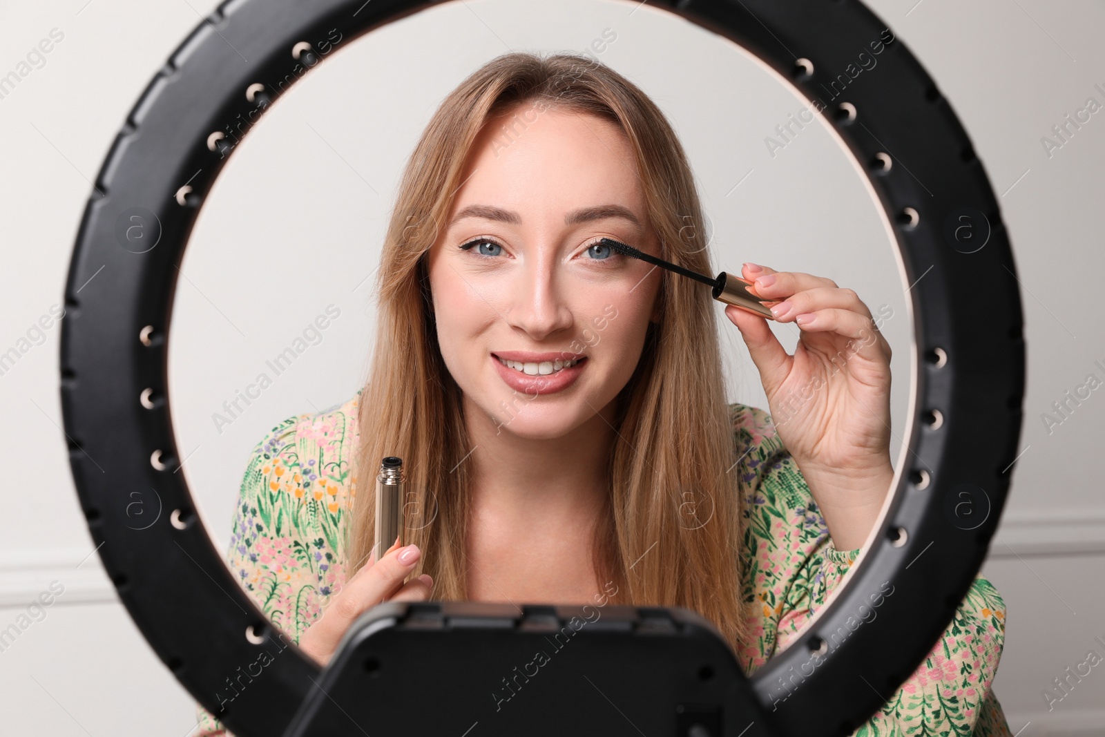 Photo of Beautiful young woman applying mascara on eyelashes near white wall, view through ring lamp