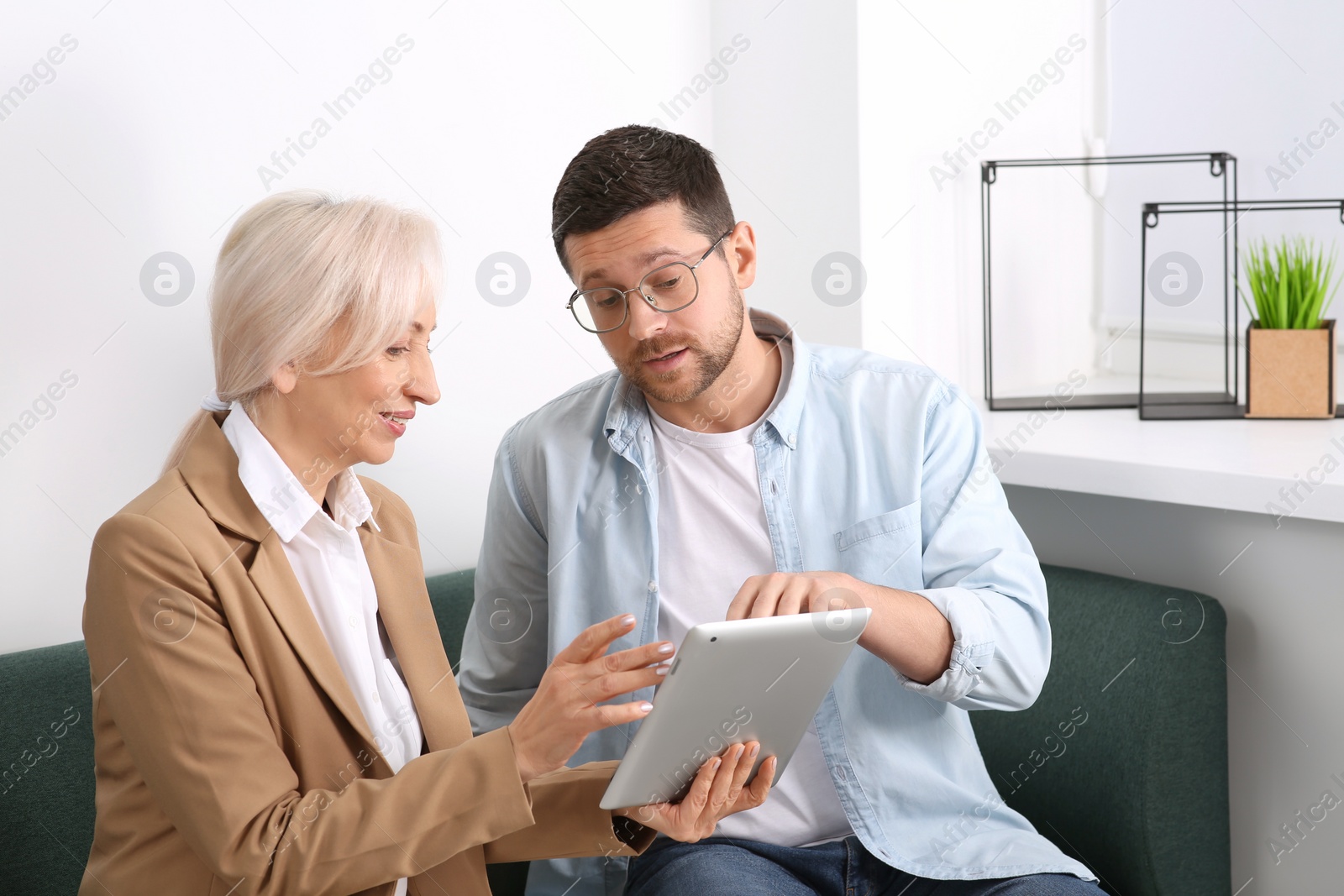 Photo of Happy boss with tablet and employee discussing work issues on sofa in office