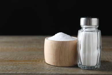 Organic salt in bowl and glass shaker on wooden table, closeup. Space for text