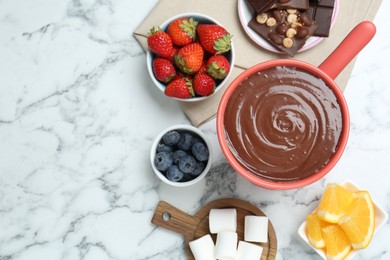 Fondue pot with melted chocolate, marshmallows, fresh orange and different berries on white marble table, flat lay. Space for text