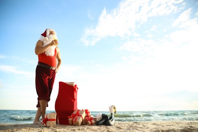 Photo of Santa Claus with cocktail and bag of presents on beach, space for text. Christmas vacation