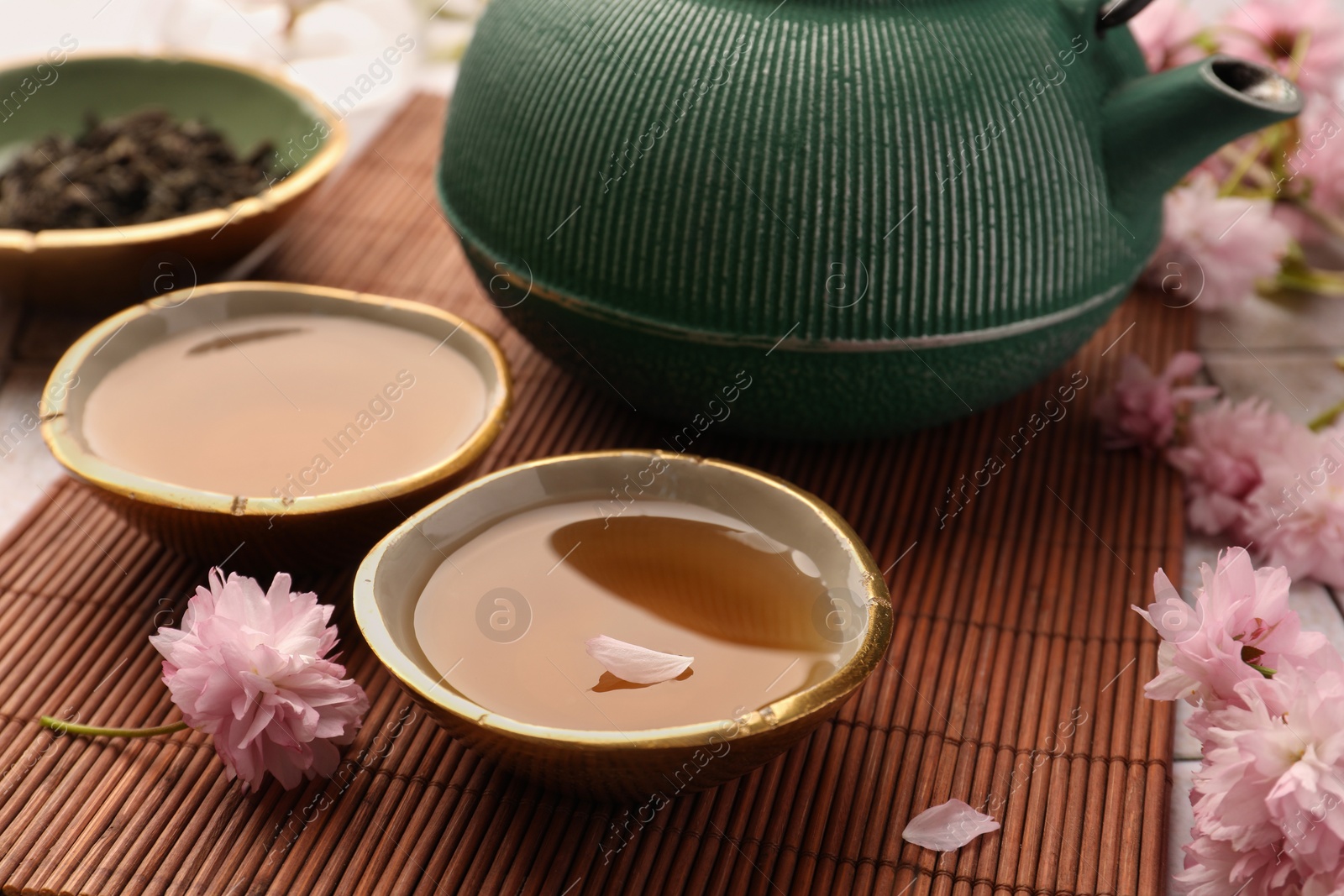 Photo of Traditional ceremony. Cup of brewed tea, teapot and sakura flowers on bamboo mat, closeup