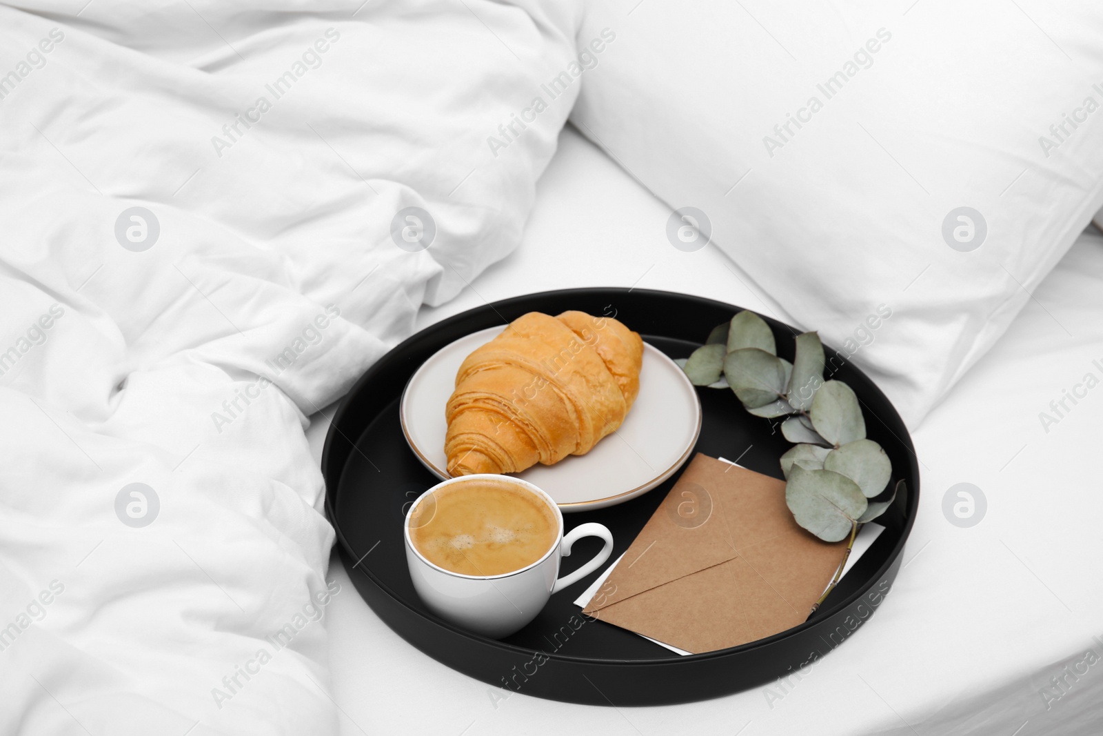 Photo of Tray with tasty croissant, cup of coffee and envelope on white bed