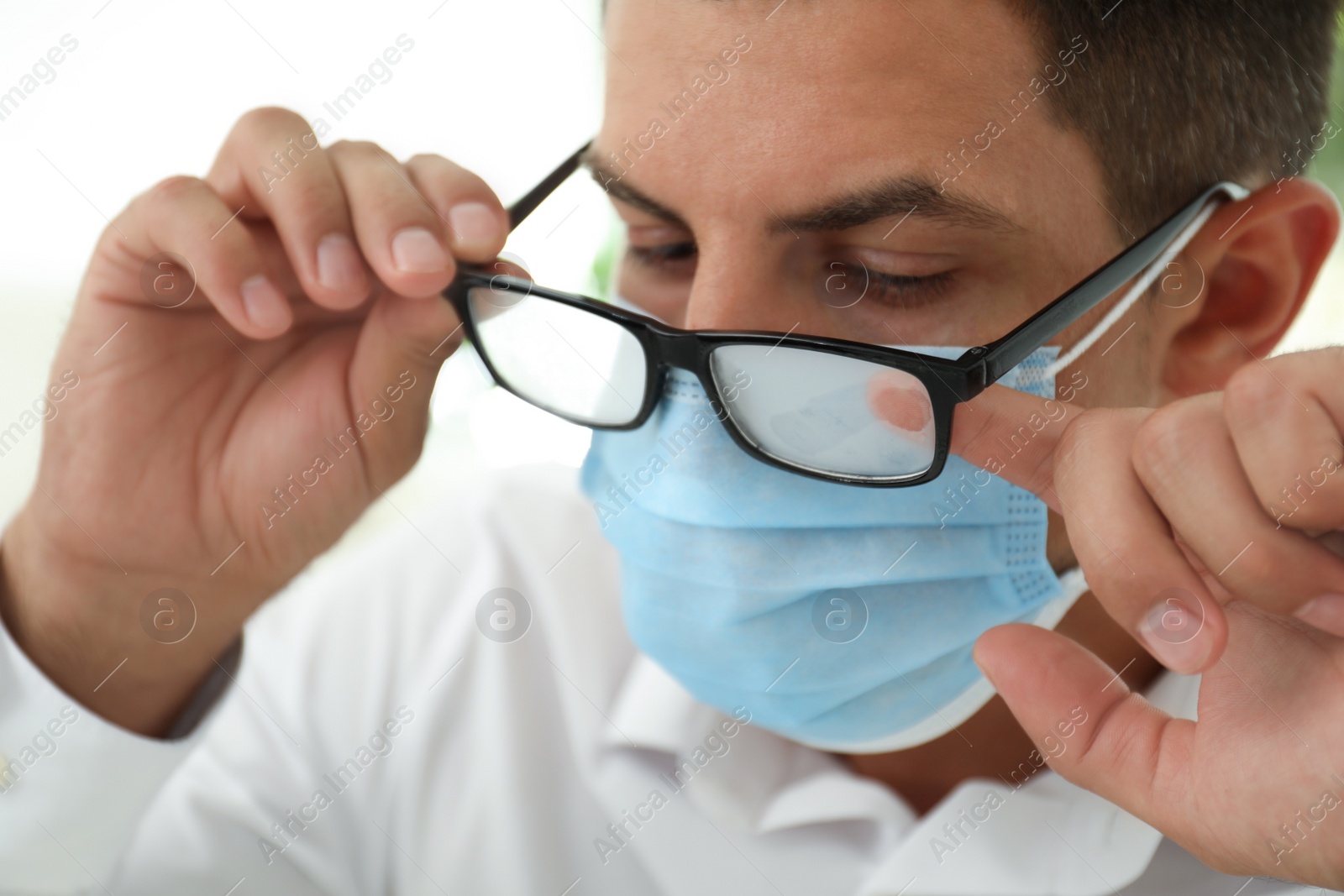 Photo of Man wiping foggy glasses caused by wearing medical mask indoors, closeup