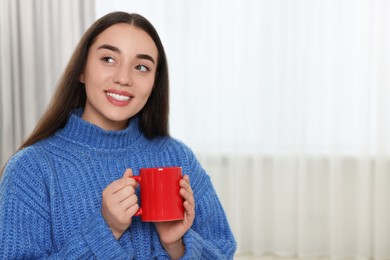 Happy young woman holding red ceramic mug at home, space for text