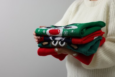Woman holding stack of different Christmas sweaters on light grey background, closeup. Space for text