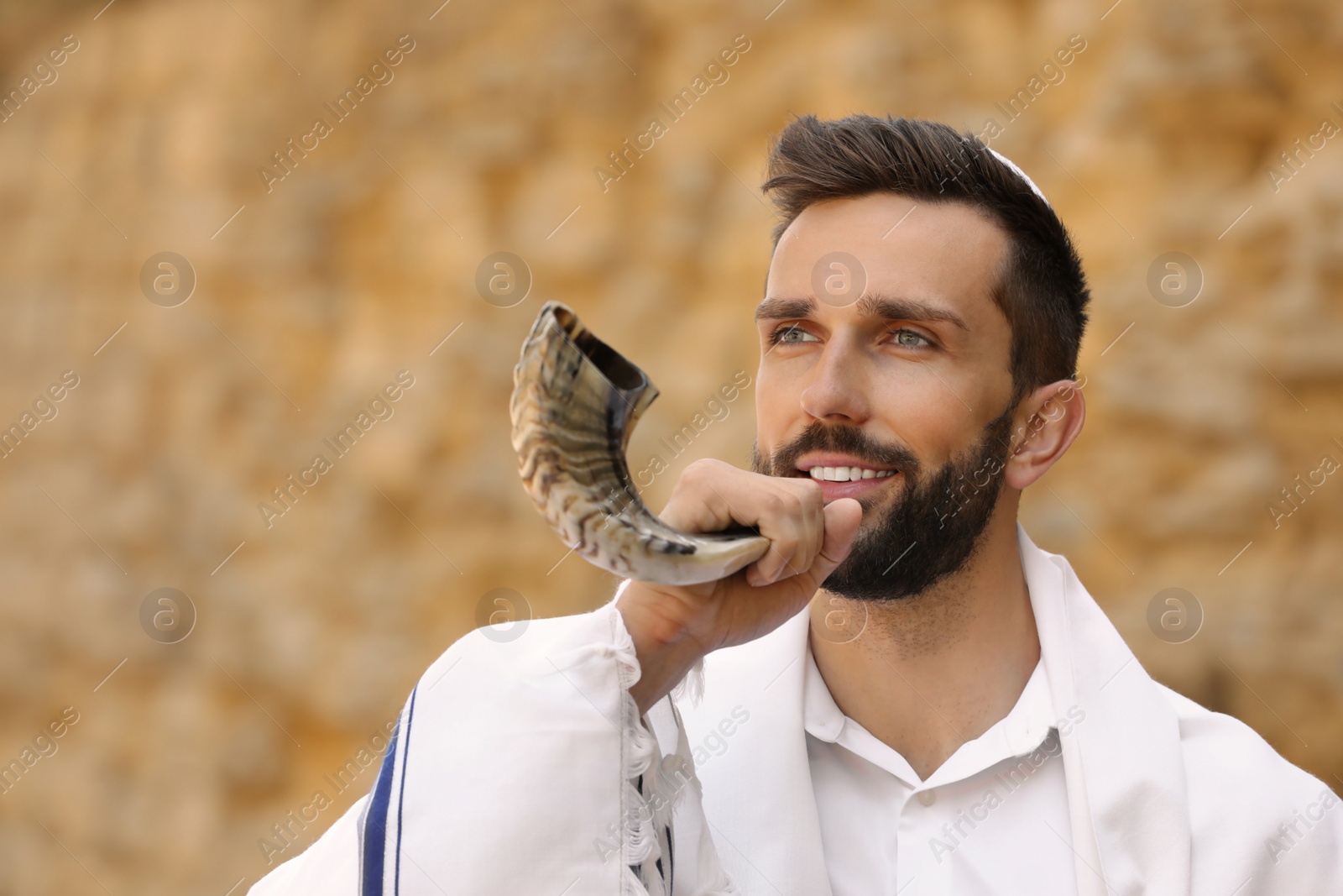 Photo of Jewish man in kippah and tallit blowing shofar outdoors. Rosh Hashanah celebration