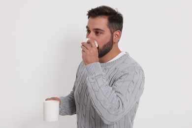 Photo of Sick man with cup of drink and tissue blowing nose on light grey background. Cold symptoms