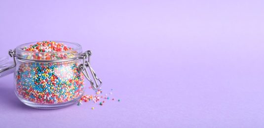 Photo of Colorful sprinkles in jar on lilac background, space for text. Confectionery decor