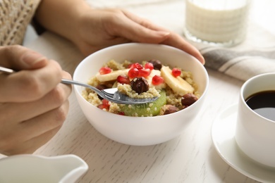 Photo of Woman eating quinoa porridge with hazelnuts, kiwi, banana and pomegranate seeds at white wooden table, closeup