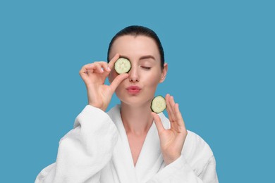 Photo of Beautiful woman covering eye with piece of cucumber and sending air kiss on light blue background