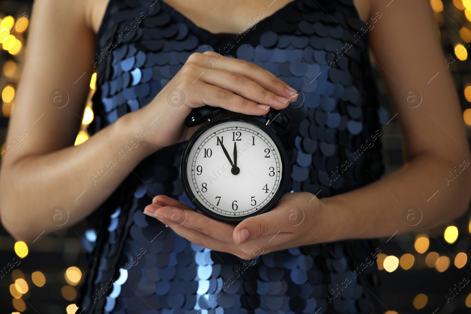 Photo of Woman holding alarm clock against blurred lights, closeup. New Year countdown